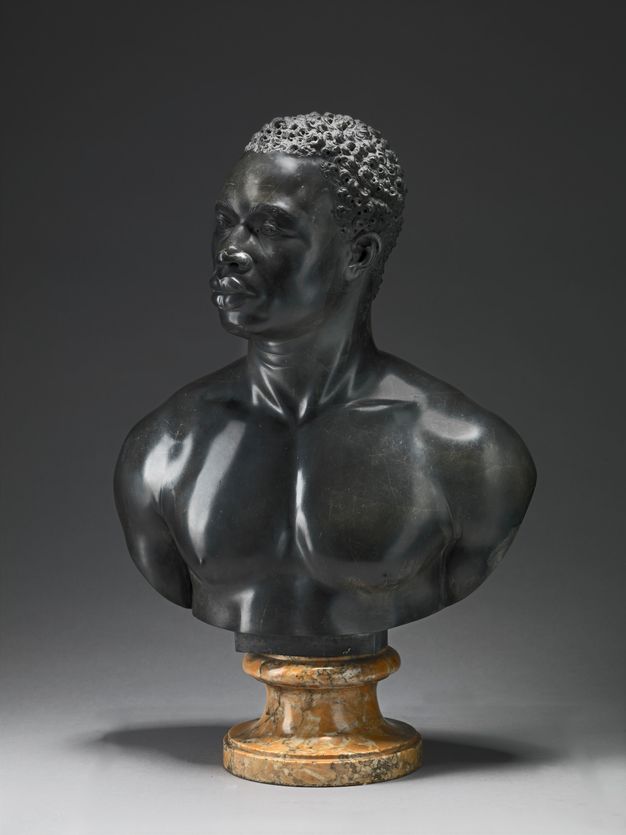 Sculpture. A bust includes the head, neck, and muscular chest and shoulders of a Black man. He has short, curly hair, noticeable eyebrow ridges, a wide nose, and his full lips are closed. Tendons stand out in in his neck, which turns halfway to look off to our left. Light gleams on the polished surfaces of the black limestome.