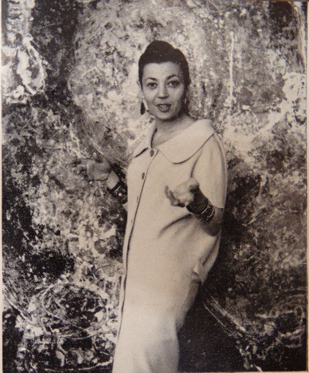 Black and white photograph. A woman, the artist Magda Cordell, stands in front of her painting Figure (Woman). The painting fills the background. Magda’s long white dress or coat has a wide, rounded collar and buttons down the front. She holds both hands up, and her lips are parted, as if about to speak or smile.