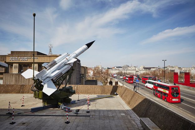 In front of a building identified on its front as 'Queen Elizabeth Hall Purcell Room', a missile launcher angles up at the sky. On a paved courtyard, the launcher is surrounded by red and white striped chains and stanchions. Several double-decker red buses drive on road running alongside the buildings to our right.