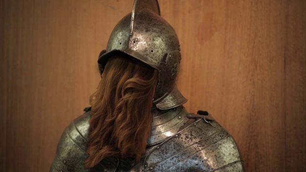 Video still. A suit of armour is shown from the shoulders up. Chestnut-brown, wavy hair pours from the opening where the face should be. The surface of the armour is darkened in areas, perhaps pitted.