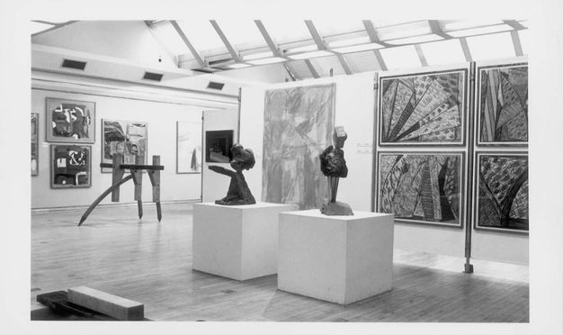 Installation photograph. This black and white photograph of two sculptures, possibly abstracted busts, on pedestals. Farther from us, another sculpture sits on pointed feet directly on the floor in a gallery hung with paintings or other media.