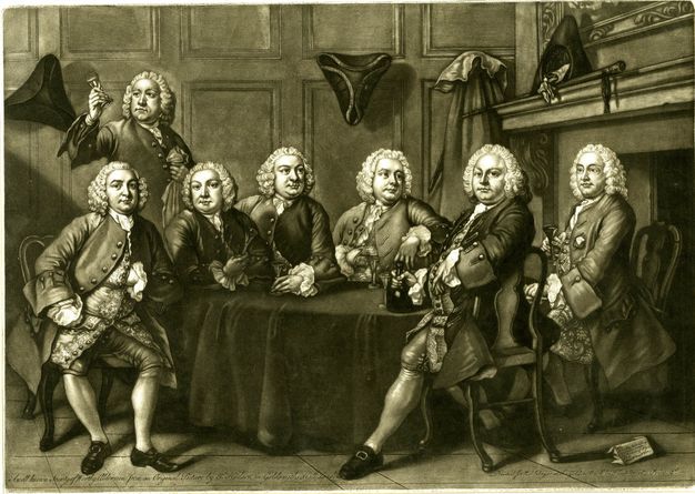 Print. Seven men with almost identical clean-shaven, pudgy, round faces sit or stand around a table in front of a hearth. They all wear white wigs and suits. Tricorn hats hang on the wall behind them or are propped up on the hearth to our right.