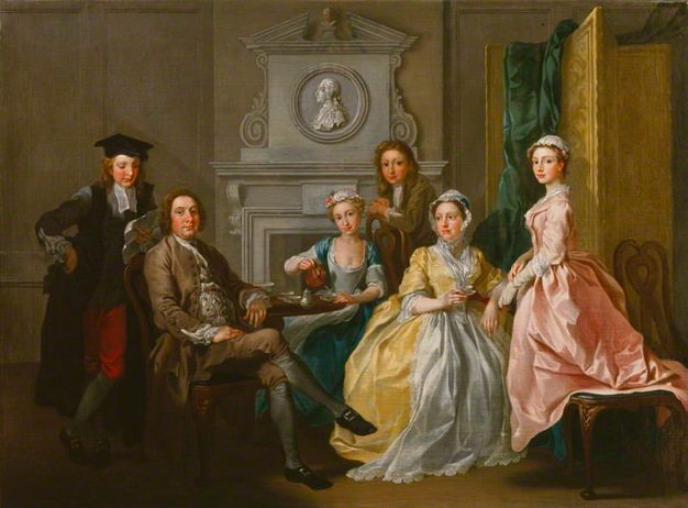 Painting. A man, woman, two younger men, and two younger women gather around a tea set on a round wooden table, placed in front of a hearth. They all have pale skin with rosy cheeks and pink lips. A profile portrait, carved like a cameo in grey, is in a medallion over the mantle.