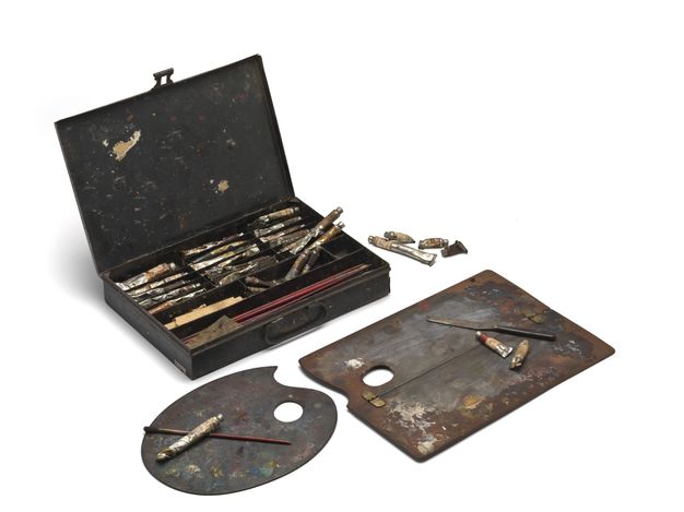 Colour photograph. A paint box holding now dented and discoloured tubes of paint and brushes is open next to one oval and one rectangular painting palette.