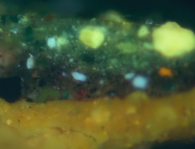 Colour photograph. In this microscopic cross section of paint, a warm, yellow layer on the bottom is topped by a green layer shot through with flecks of green and blue.