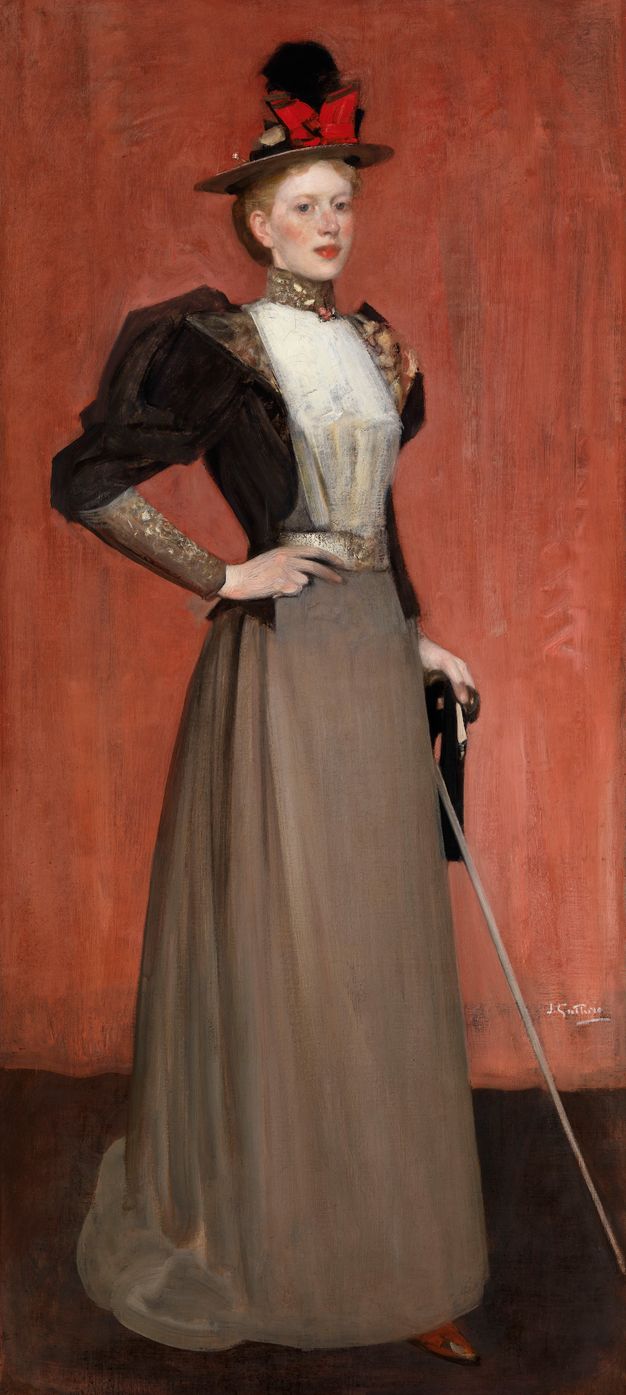 Painting. A woman wearing an elegant dress and hat stands in front of a brick-red curtain, looking out at us in this tall portrait. The woman has pale skin, flushed cheeks, and red lips. Her brimmed hat has a red bow and black object, possibly a feather. The high neck of her dress is gold, and her shirt crisp white against a fitted black jacket with puffed sleeves. Her smoke-grey skirt falls smoothly to the floor. One hand is on her hip, the other holds a cane.