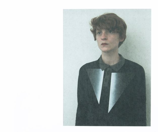Colour photograph. Shown from the waist up, a person with pale skin and a mop of brown hair looks up and to our left with dark eyes. They have a nose ring, and full, red lips are closed. Below their black shirt’s collar two triangular pieces extend far down the chest like exaggerated lapels. They seem to be flat and shiny, as they deepen from white along the middle to black on the outer corners.