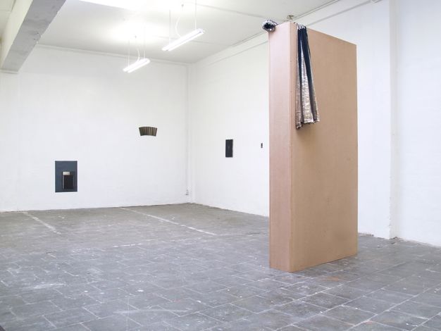 Installation photograph. Four works, all in shades of blue or brown, hang sparsely on white gallery walls. A grey stone floor looks worn and possibly spattered with paint. To our right, a navy blue and silver cloth hangs over a narrow floating wall, which reaches close to the ceiling.