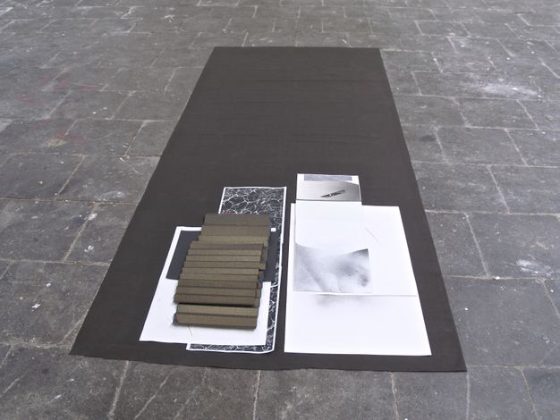 Installation photograph. Several works on paper are layered at one end of a long, rectangular black cloth laid on a grey stone floor. One sheet appears rather large, perhaps A2 size paper. Others are long strips or smaller pieces. Forms and patterns on the papers are in black, white, and shades of grey. Two golden brown pieces are folded like accordions.