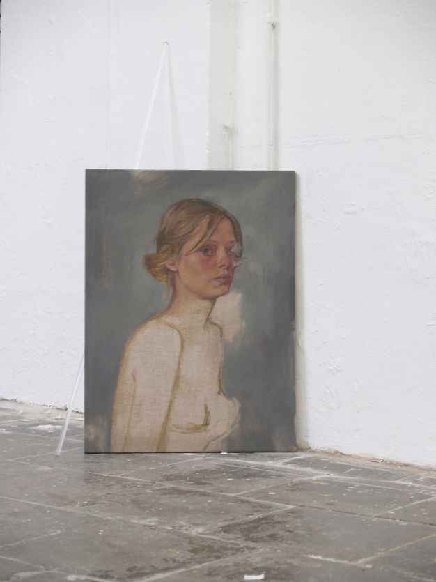 Installation photograph. A painting shows the head and shoulders of a pale-skinned woman with dark blond hair. Her face and hair are painted in detail. Bare linen is left where her body, including breasts, would be. The background is elephant grey. The painting sits at an angle on the floor against a white wall of a gallery. Behind it, a clear plastic rod leans against the wall.