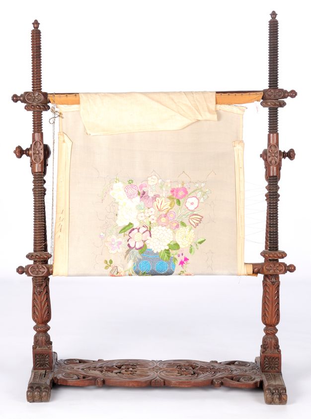 Embroidery. A strip of cream-white cloth stretched on a wooden stand is embroidered with a vase of flowers. Some of the flowers have pink, yellow, red, and green. Others are blocked out but not yet stitched, or seem to be sketched onto the cloth. The cloth is rolled over dowels in the carved frame, and the height between the rails can be adjusted on either side.