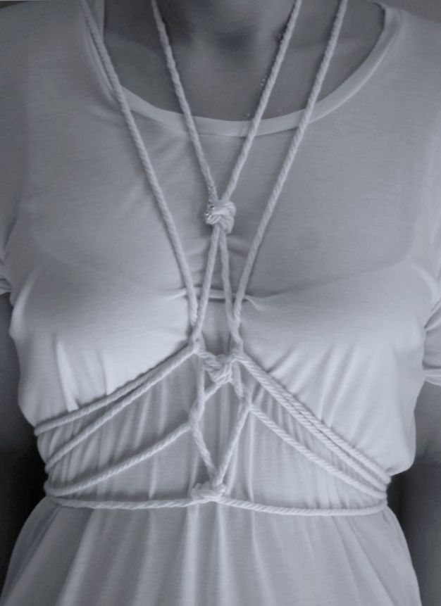 Black and white photograph. A woman’s torso is tied with intricately knotted white rope over a white shirt. The contours of her bra are visible through the thin fabric.