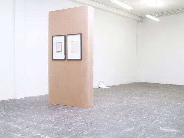 Installation photograph. Two framed works hang on a narrow floating wood wall in a gallery with white walls and a grey stone floor. The frames are black and the works within nested rectangles in shades of grey and white.