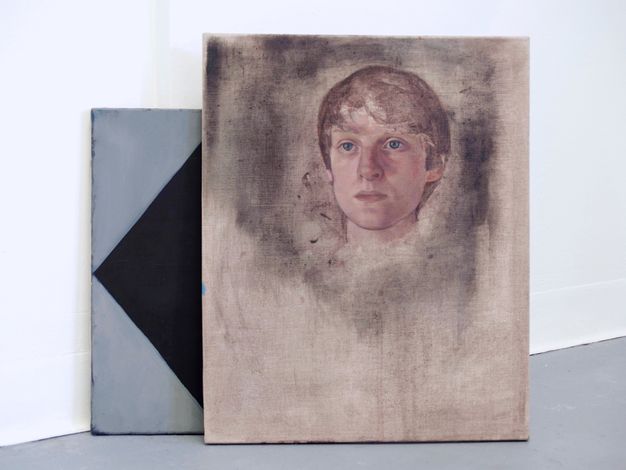 Installation photograph. Two works lean against the white wall of a gallery. The front painting is of a pale face gazing steadily off to our left with blue eyes. Straight brown hair falls in bangs and is cut close to the head. A dark grey area around the head is thinly applied, as if rubbed into the linen. The work tucked behind it is a design of grey and black triangles.