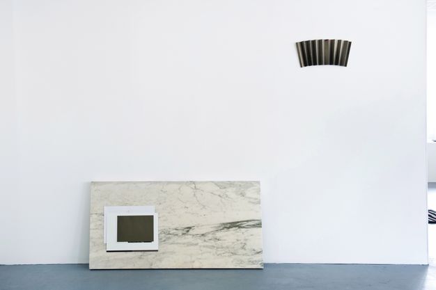 Installation photograph. A panel resembling marble rests on a grey floor and leans against the white wall of a gallery. Two or three black or white panels, perhaps pieces of paper, are layered on the panel. A piece of accordion-folded dark paper hangs on the wall to our right.