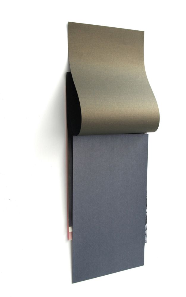 Installation photograph. A charcoal-grey piece of paper hangs on a white wall. A burnished gold-coloured sheet hangs from the wall above the grey, as if the sheets were glued together along the top like a notepad. Slivers of black, white, and pink paper peek out from under the grey sheet.