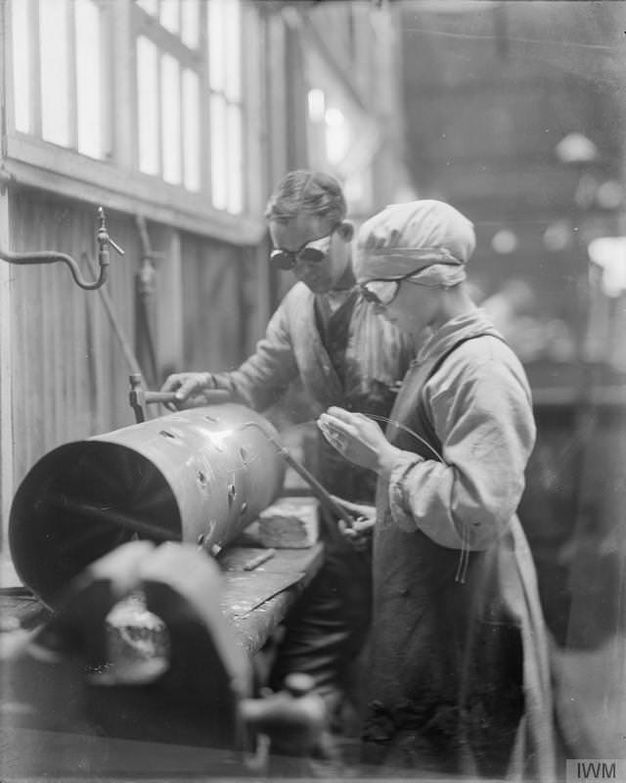 Black and white photograph. A man and young woman weld a long, metal tube in a factory. The woman wears a white cap covering her hair, and both wear reflective silver goggles.