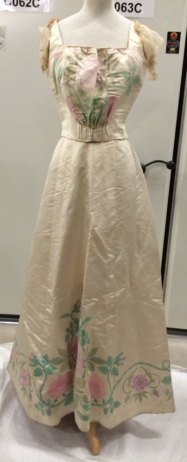 Clothing. The short-sleeved bodice of this dress has a square neckline. The skirt falls in an A-line to the floor. The fabric is ivory-white satin so has a sheen. Pink flowers and green stems and leaves are painted on the front of the bodice and around the bottom hem of the skirt.