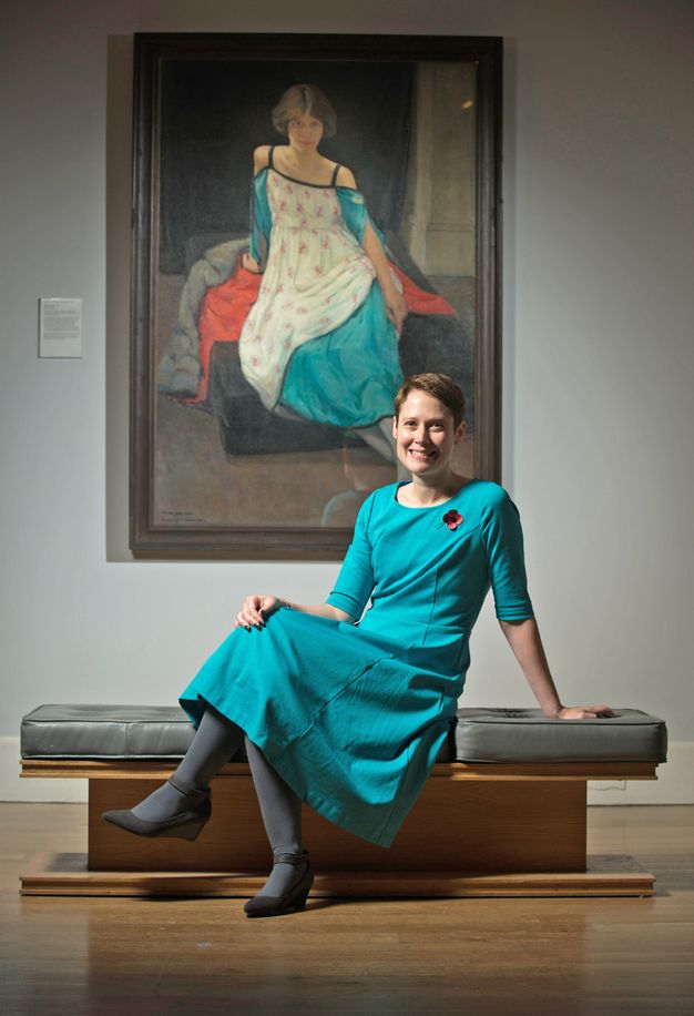 Colour photograph. A woman with short brown hair sits on a bench in a gallery and smiles at us. Behind her, a painting shows a woman who sits in a similar but mirrored pose. They both have pale skin, both look out at us, and wear azure-blue dresses.