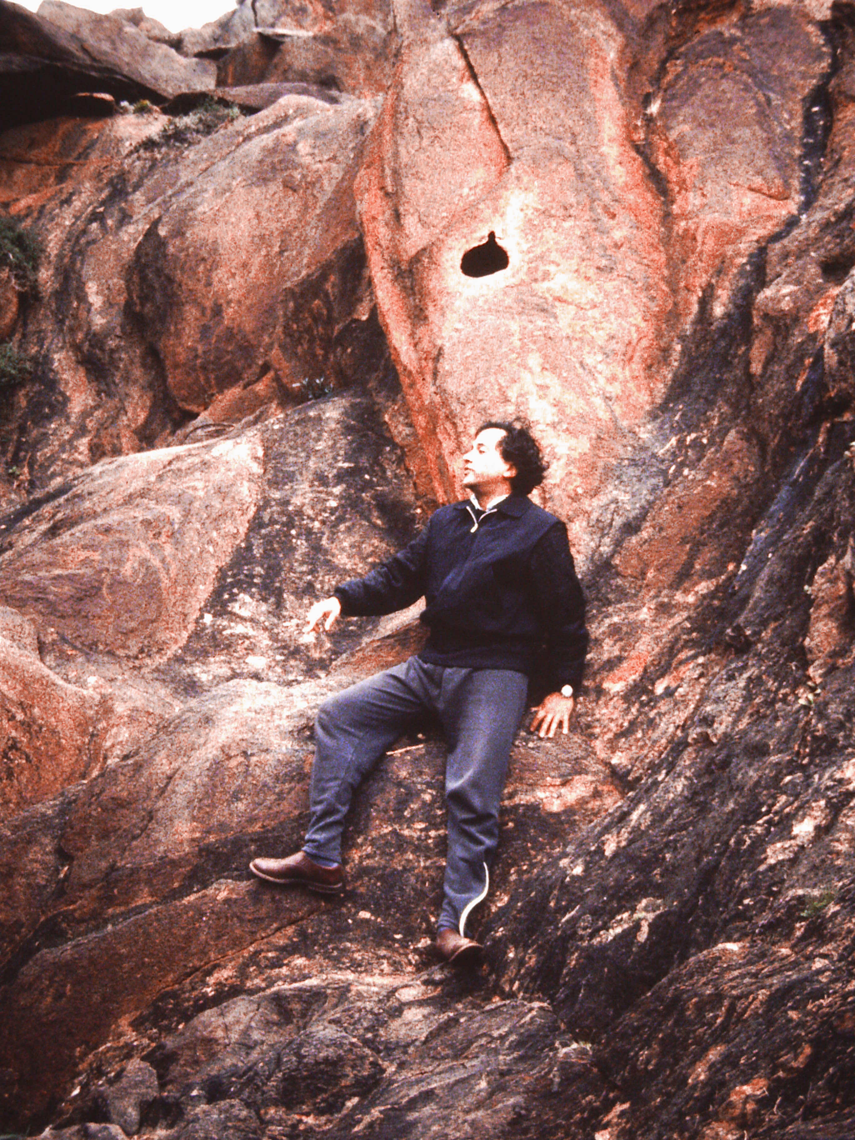 A man lies against red rock striated with black. He turns his face in profile to our left under a dark hole in the rock above his head.