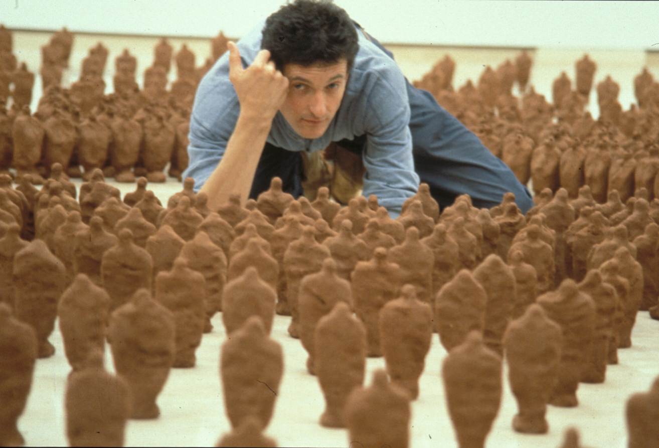 Installation. An artist crouches low to the ground and looks out over hundreds of roughly human-shaped figures. Each statuette is just over twenty centimetres high.