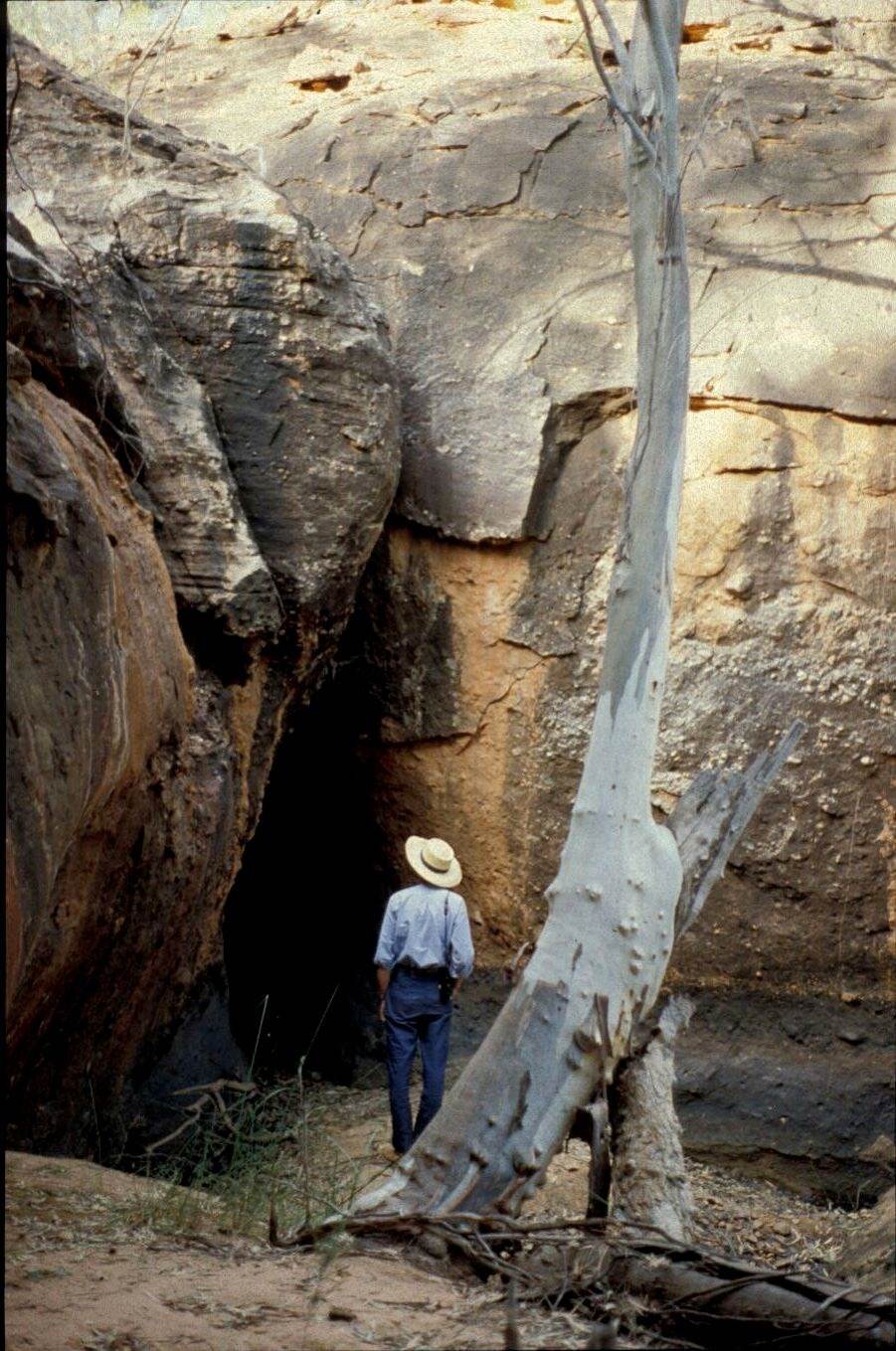 A man wearing a broad-brimmed hat stands facing away from us, looking towards the upside-down, narrow V of a cave entrance. He comes just over halfway up the height of opening. The stone face of the cave fills the picture.