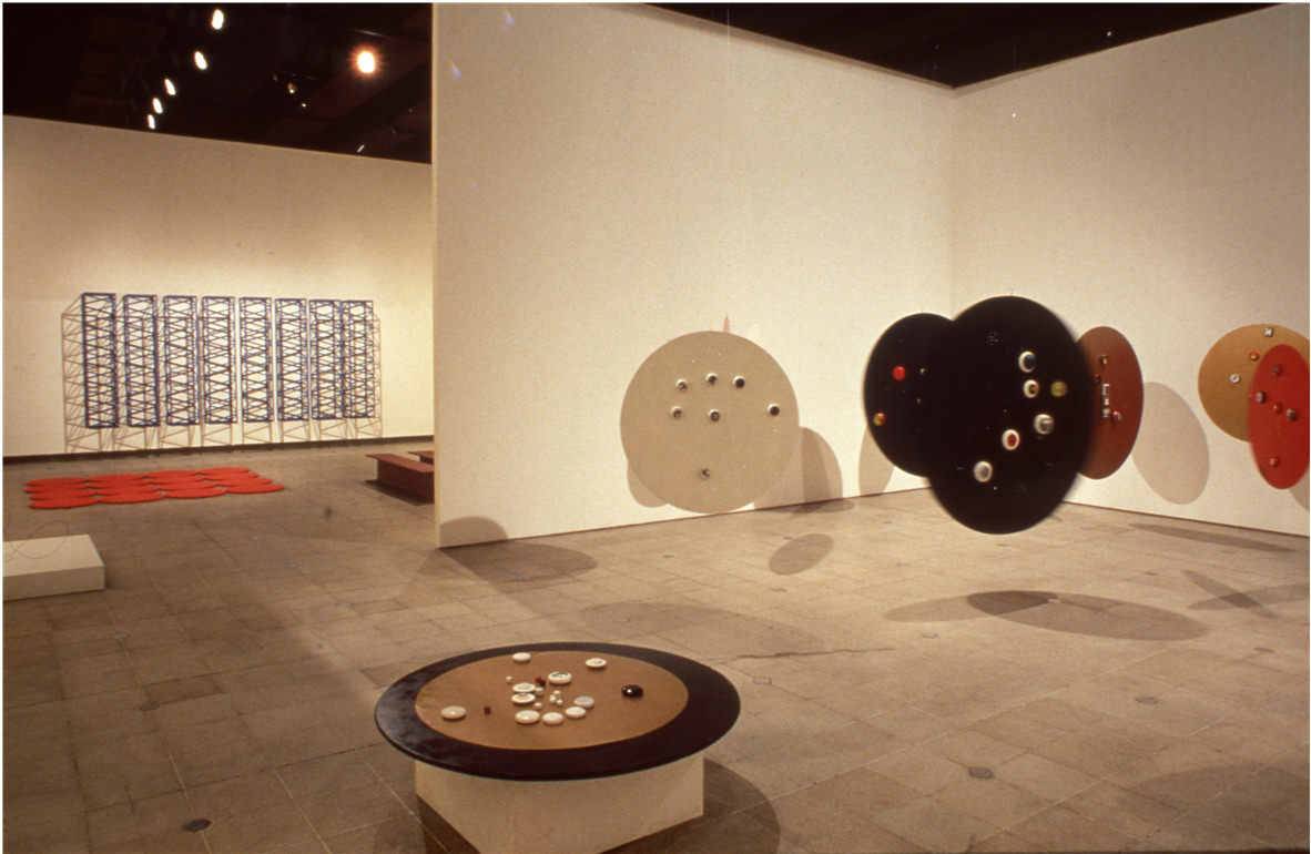 Installation photograph. Six round discs, like tabletops, float in a gallery space, presumably suspended from the ceiling, except for one, which sits on a pedestal. The discs are painted brown, red, or black.