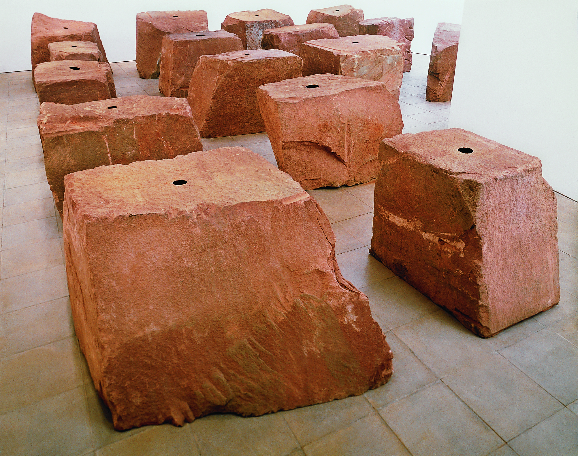 Sculpture. Sixteen roughly cube or rectangular red sandstone blocks sit in rows in a gallery. Each has a black circle at the top centre.