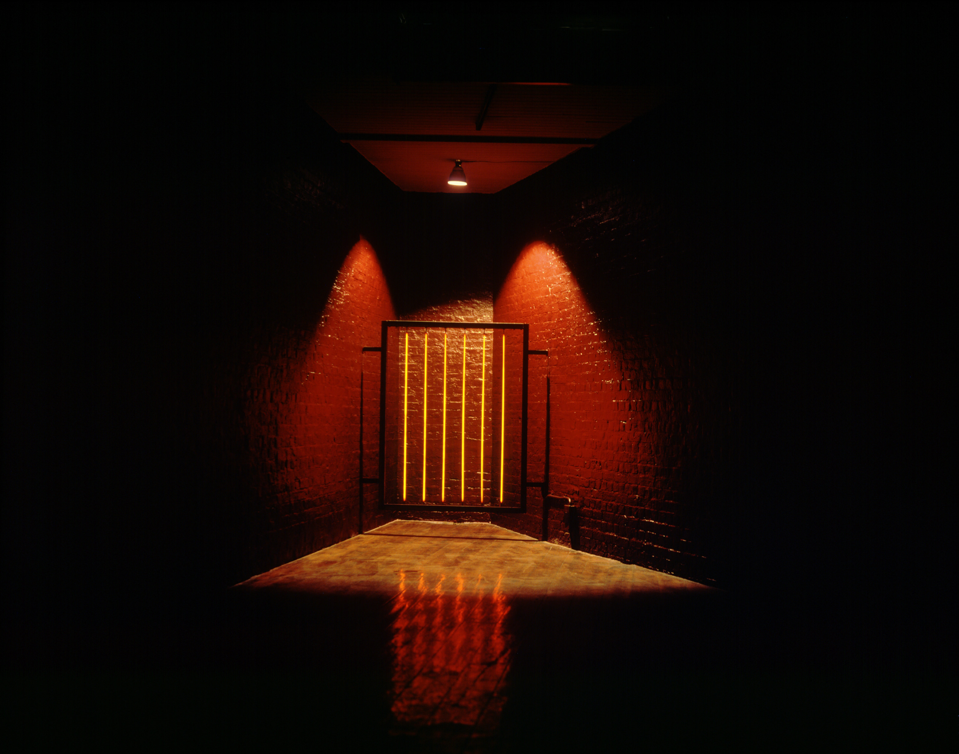 Installation. In an otherwise dark room, a spotlight creates a pool of light around a gate-like sculpture tucked into the corner of brick room, which appears to be painted red. The vertical bars of the sculpture are yellow and the framework black.