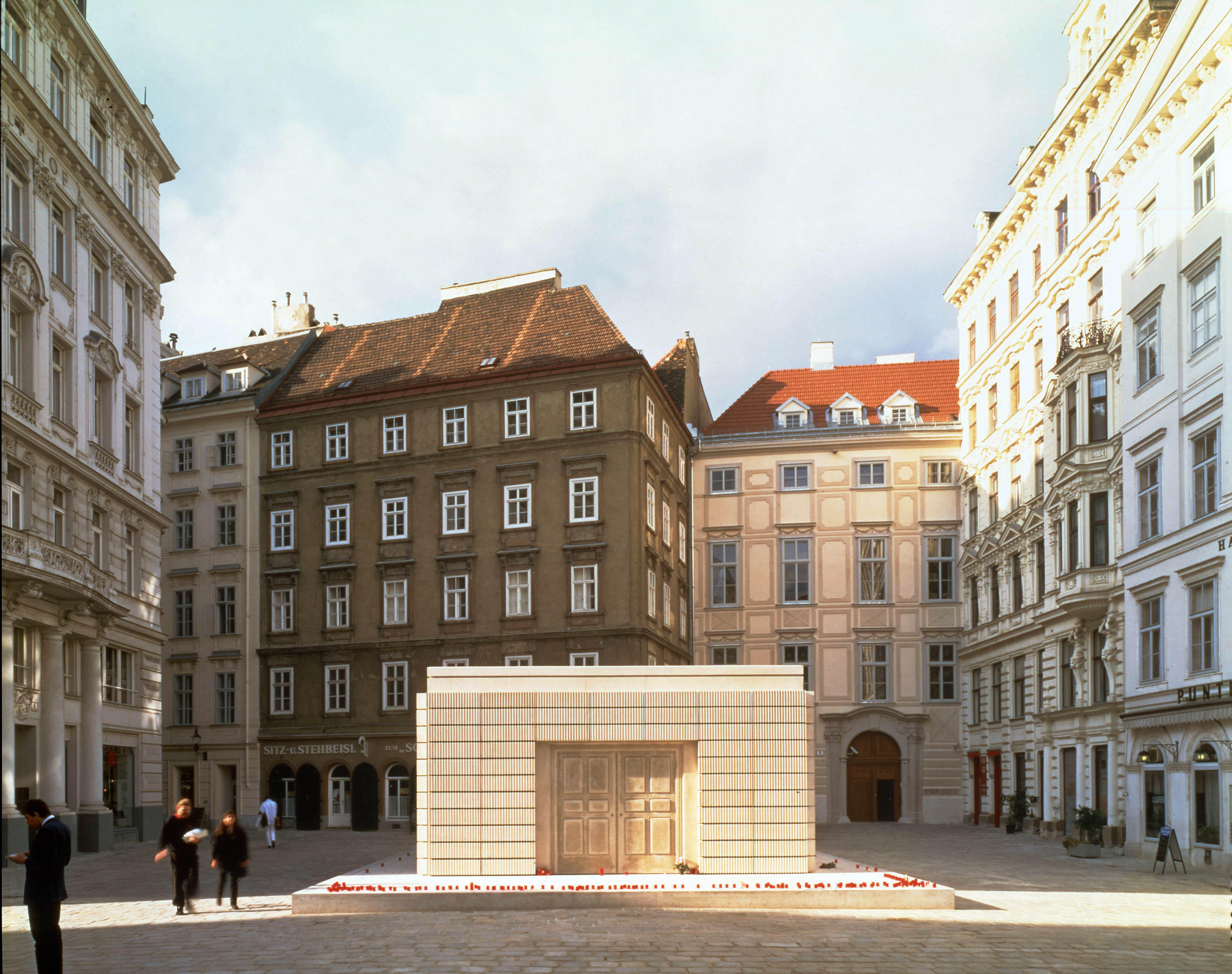 Sculpture. A two-storey, concrete cast of the inside of a library creates a sculpture capturing the negative space of the original room. Five or six-storey buildings surround the town square where this is displayed.