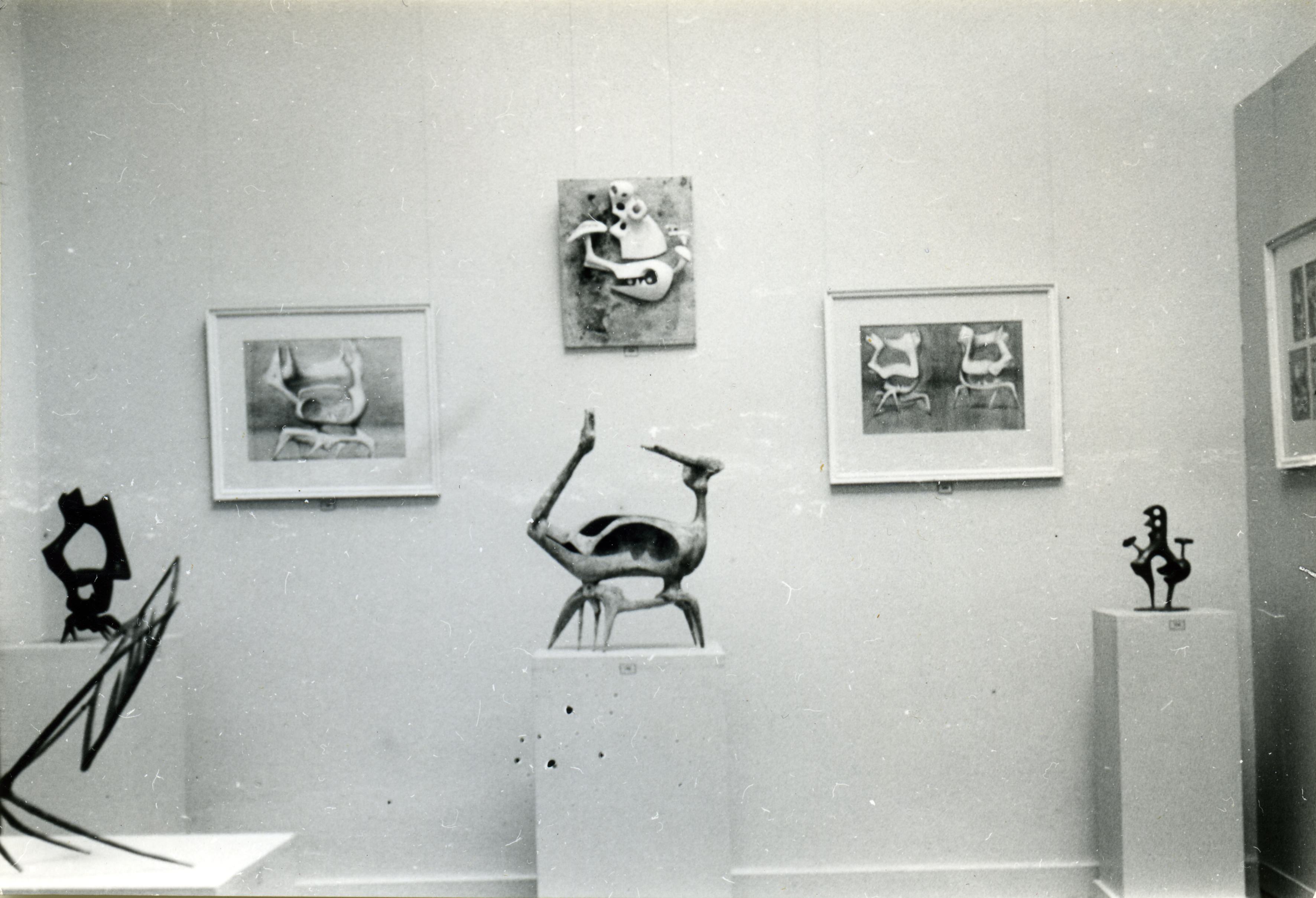 Installation photograph. This black and white photograph shows four sculptures on plinths in front of a gallery wall hung with three abstract works. The middle of the hanging works has shapes in relief against a flat background.