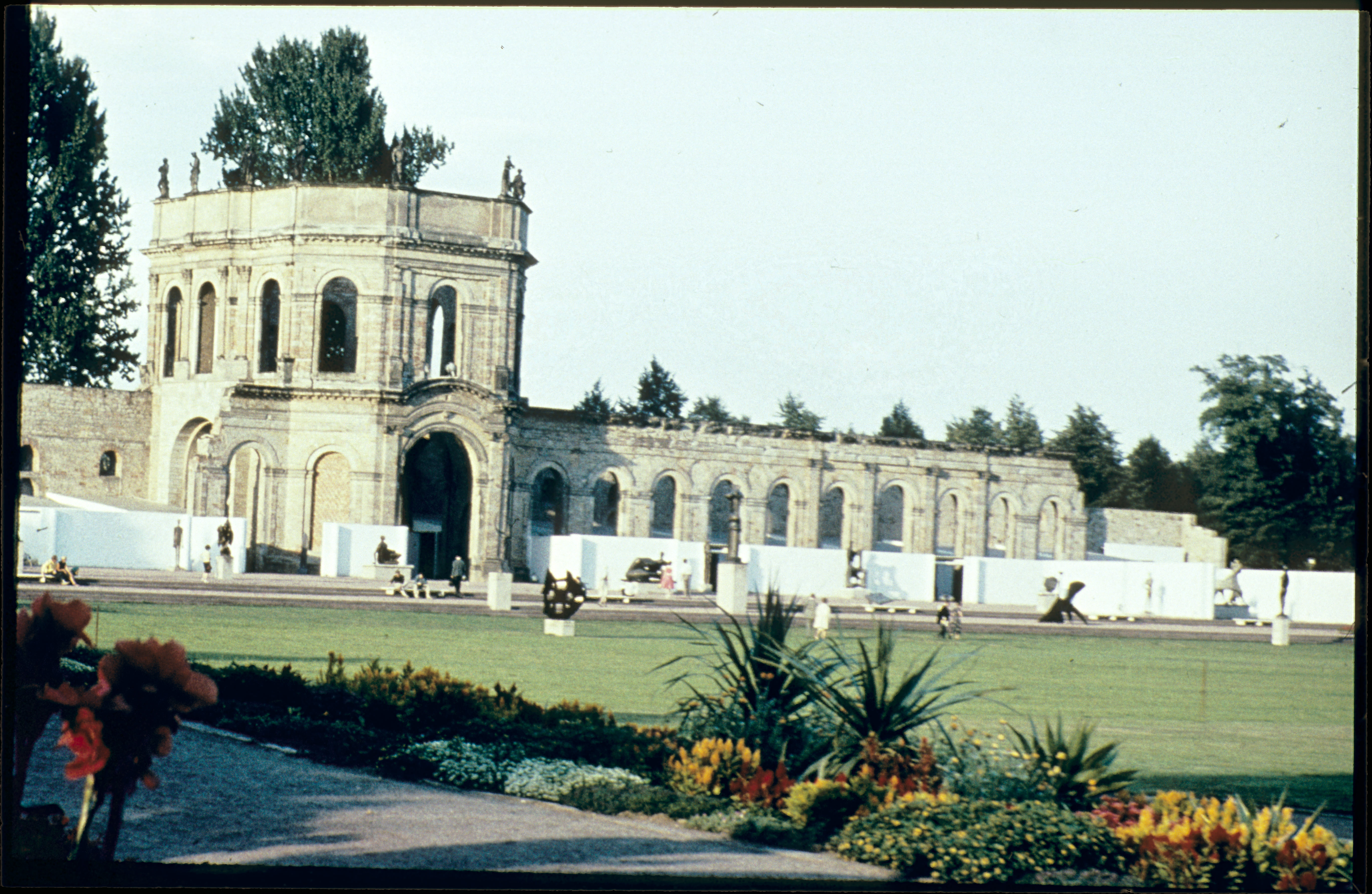 Installation photograph. Abstract and stylized sculptures are displayed on plinths and in an open-air stage set up at the far side of an expanse of grassy lawn. The stage spans the length of a colonnaded stone conservatory.