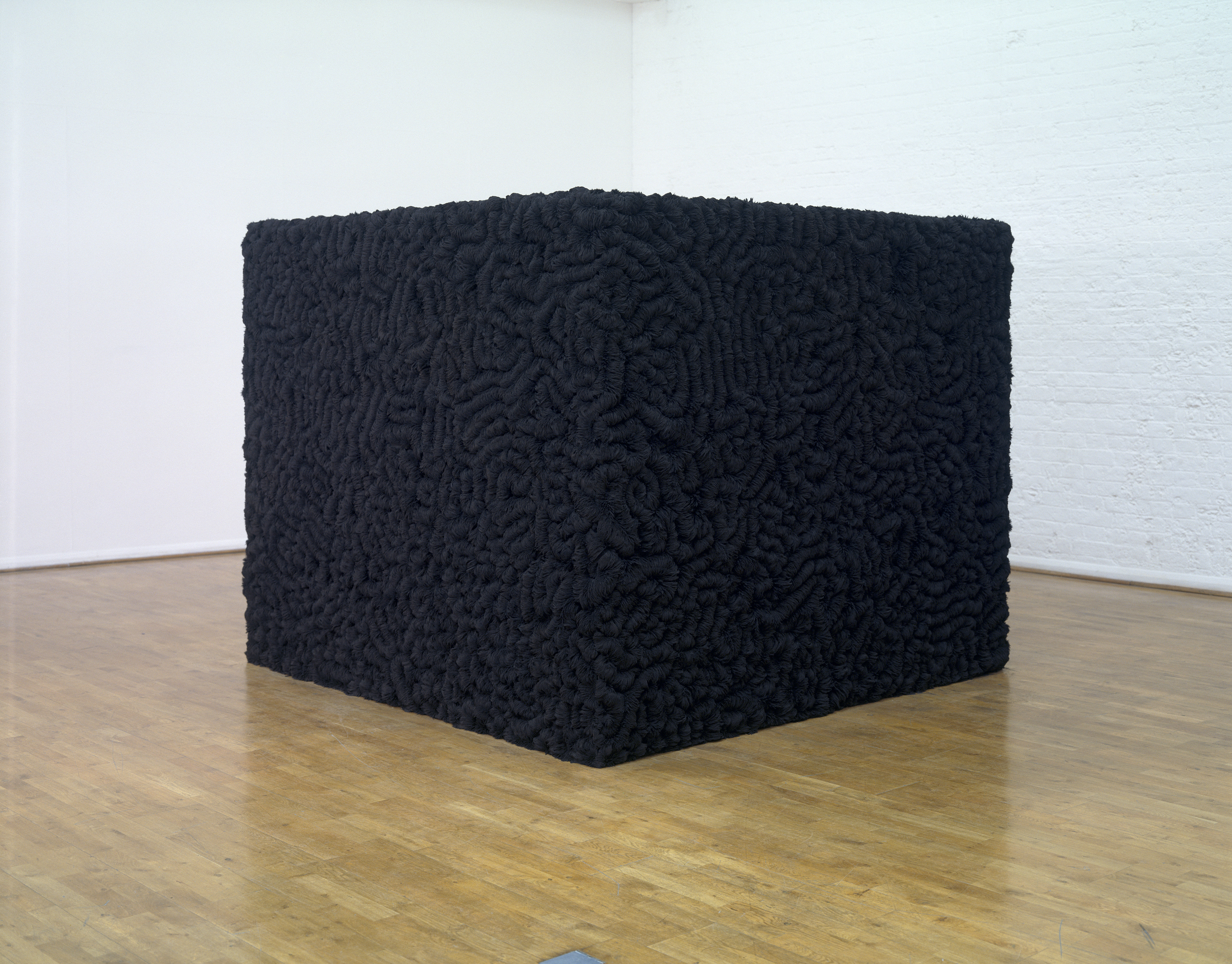 Sculpture. A black box sits near the corner of a gallery. Every visible surface is covered with dense folds, like a human brain.
