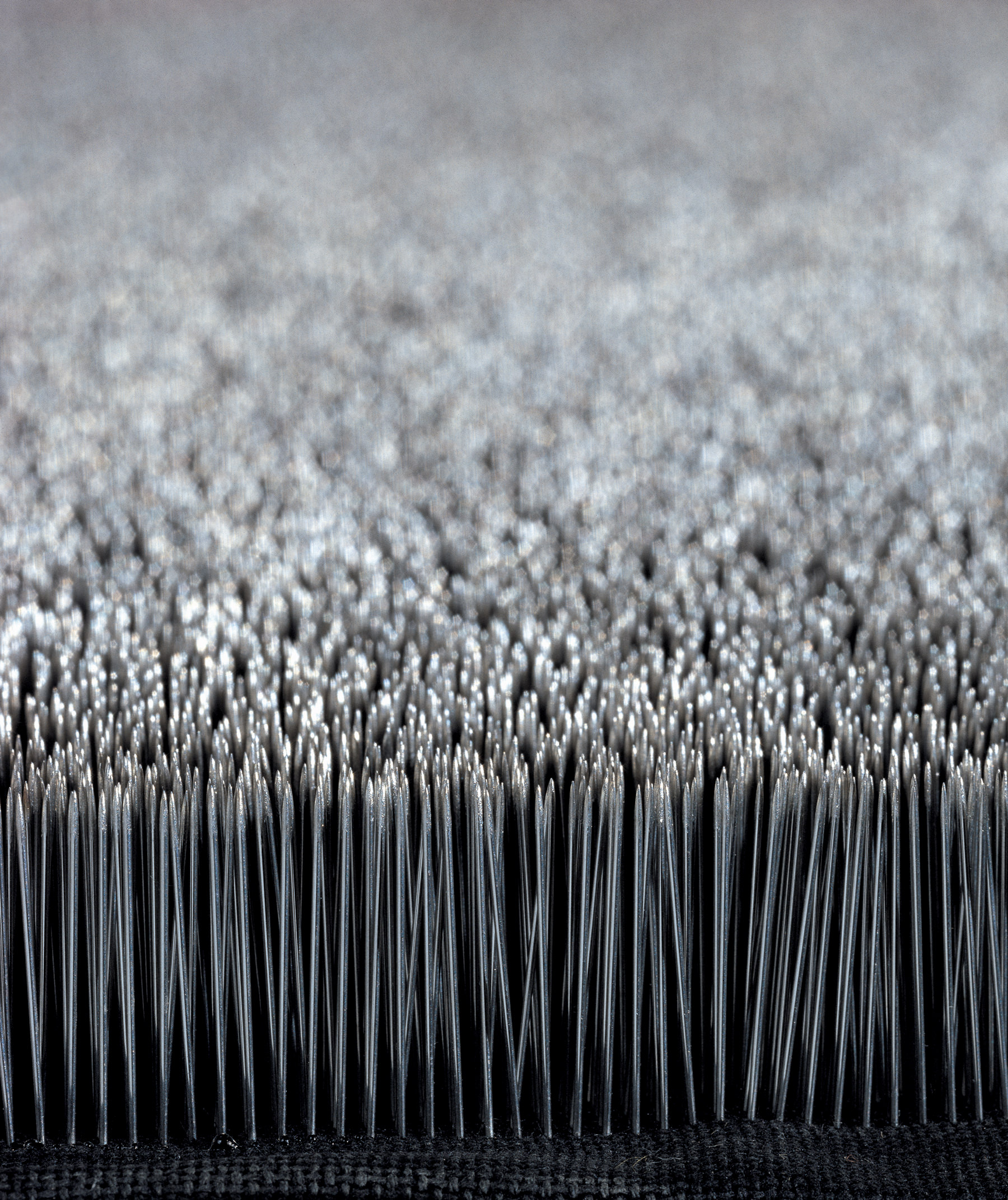 Sculpture. Detail view of rows of countless pins, set so their points face upwards. The surface created by the pinpoints shimmers silver.