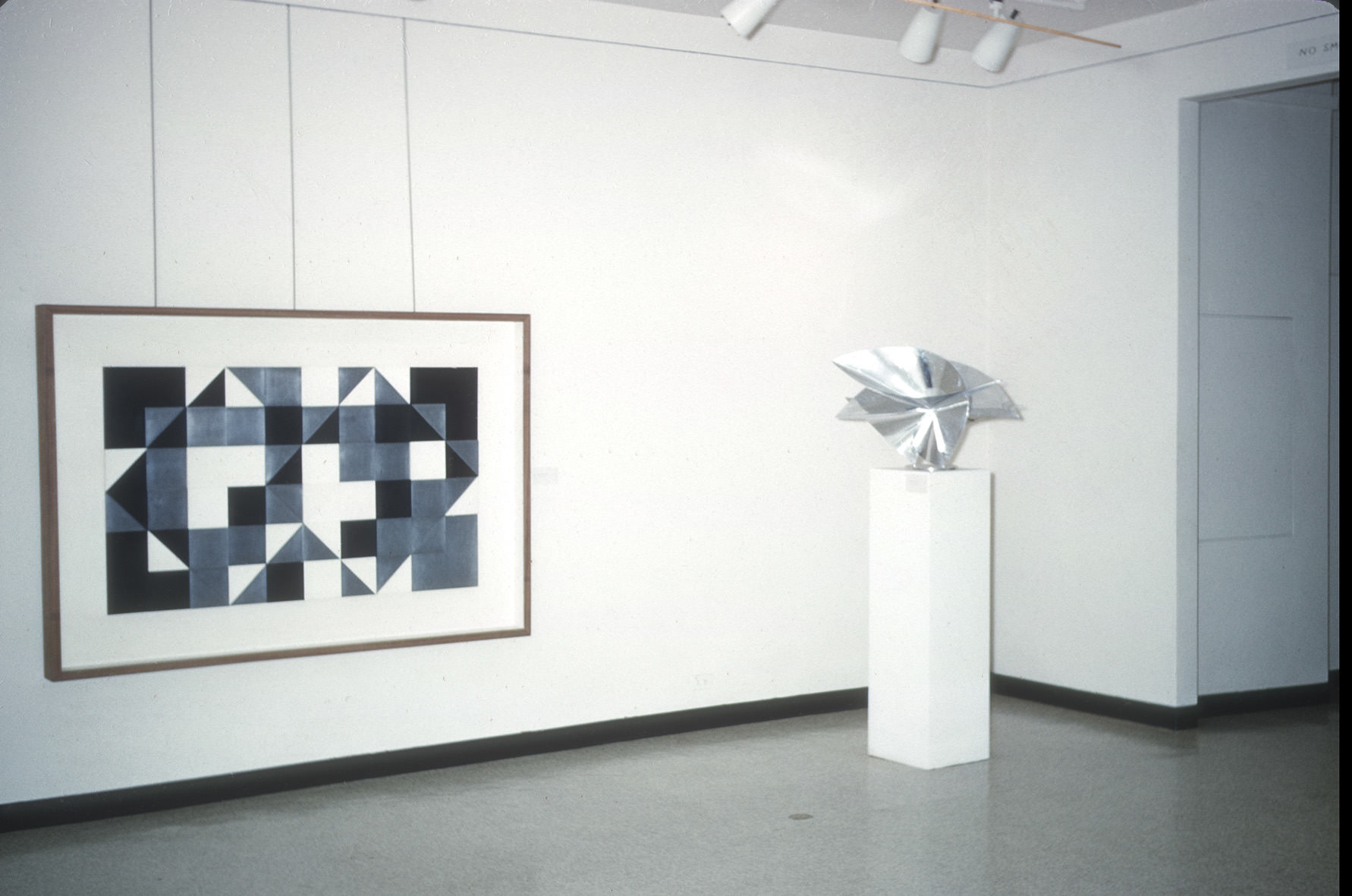 A gallery space with a framed geometric artwork on the left and a metallic sculpture on the right. The artwork features a pattern of interlocking black, white, and gray triangles and squares, creating a bold, abstract design. The sculpture, displayed on a white pedestal, is a shiny, angular form with folded and curved elements, giving it a dynamic, reflective appearance. The background consists of clean, white walls with minimal decor, emphasizing the art pieces.