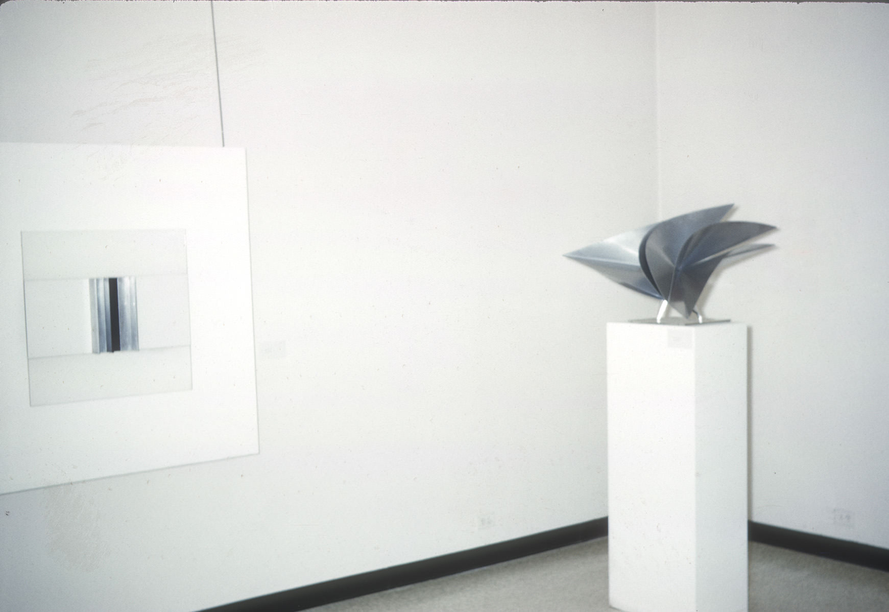 A gallery space featuring a minimalist artwork and a sculptural piece. On the left is a framed artwork with a white background and a central vertical element composed of reflective metallic or glass material, creating a sleek and modern aesthetic. On the right is a metallic sculpture displayed on a tall white pedestal. The sculpture consists of curved, intersecting, and pointed forms, showcasing a dynamic and abstract design. The clean, white walls and sparse layout emphasize the contemporary nature of the pieces.