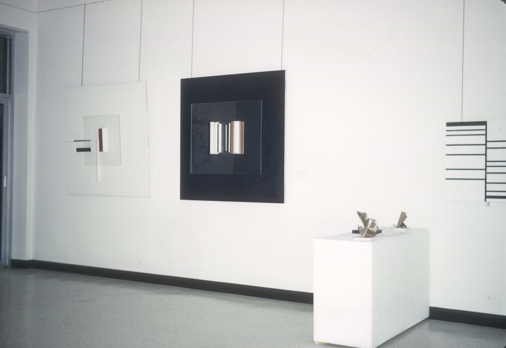 A gallery display featuring three framed minimalist artworks on the wall and a pedestal with small sculptural pieces. The left artwork is a translucent, layered composition with abstract geometric elements in black, white, and red. The center artwork is framed with a black square and features vertical bars in metallic and neutral tones, creating a reflective effect. The right artwork is composed of black horizontal lines arranged in a grid-like structure on a white background. In front of the artworks, a white pedestal holds small, abstract sculptures with metallic finishes, showcasing sharp and angular designs. The clean and bright gallery space enhances the modern aesthetic.