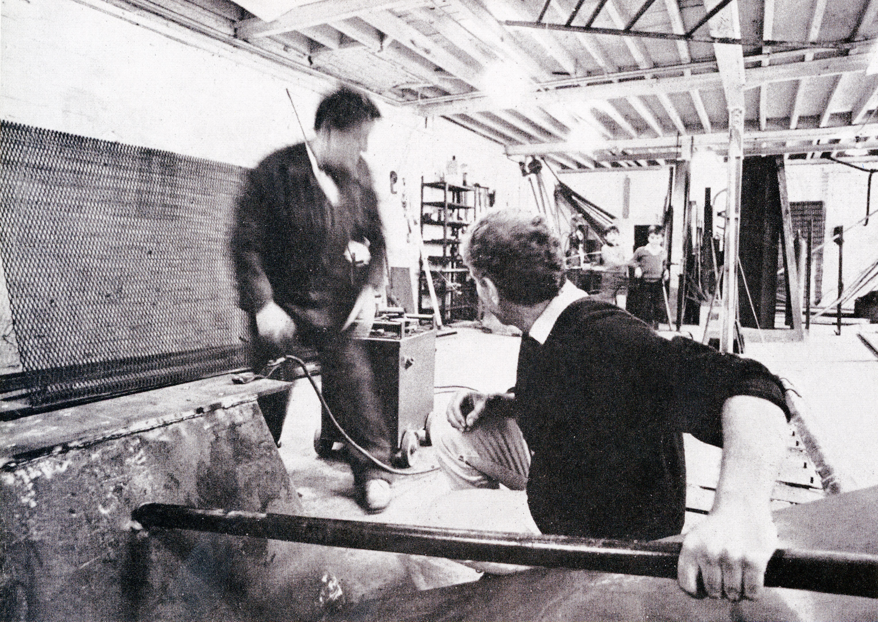 Black and white photograph. Two men work in a studio. One grips a long metal tube and both turn to look at two children standing in the background.