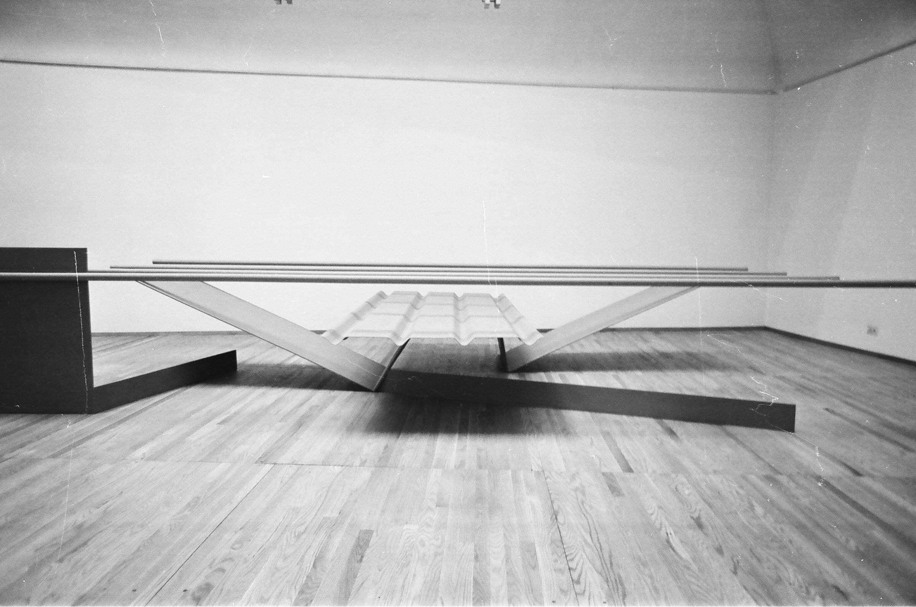 Installation photograph. Black and white photograph of an abstract sculpture in a gallery. Four tubes balance across an arrangement of V-shaped metal panels.
