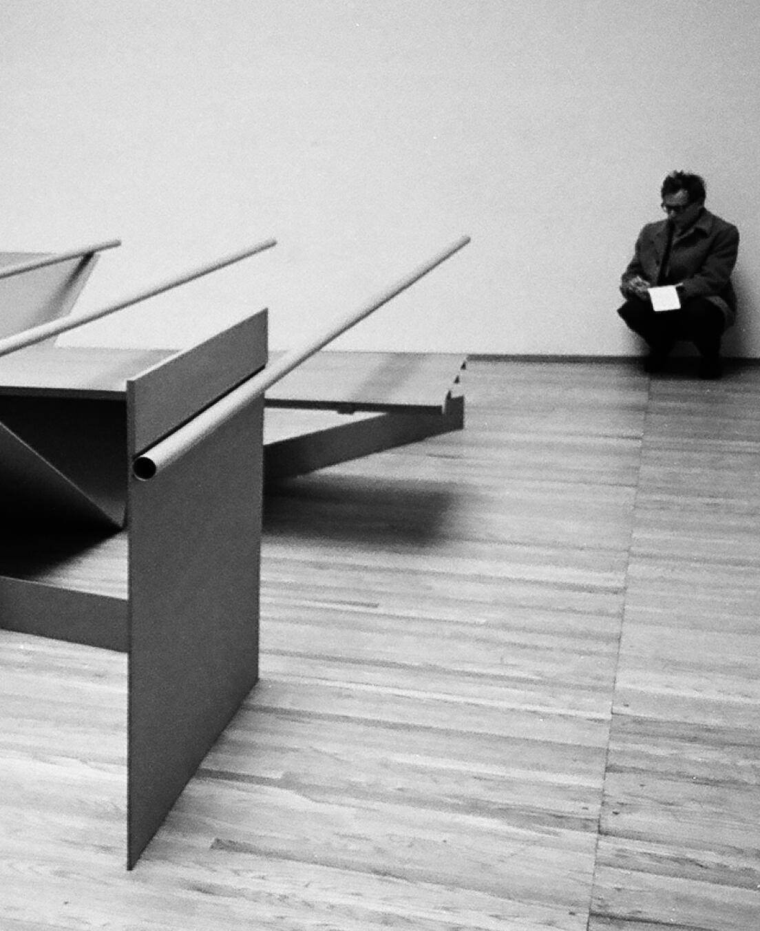 Installation photograph. Black and white photograph of the edge of an abstract sculpture. A long metal tube affixed to a vertical panel to at one end, and the other seems to float. A man crouches against the far wall.