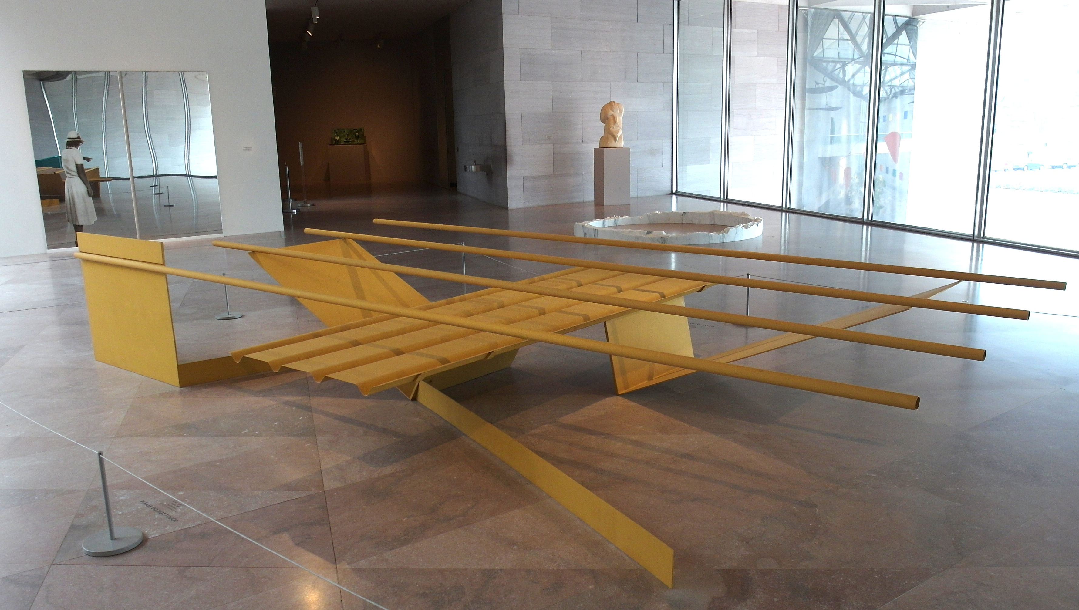 Installation photograph. An abstract yellow sculpture made of four tubes balanced across vertical, horizontal, and V-shaped metal panels is displayed in a museum with pink marble floors. Other sculptures are exhibited in the background.