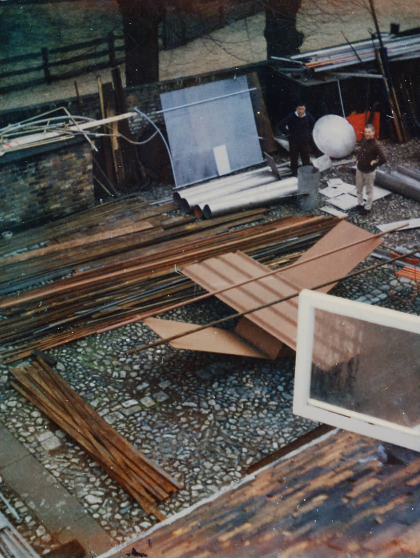 Colour photograph. We look down into a courtyard where a tawny-brown, abstract sculpture is set up. Two long tubes appear balanced across flat and V-shaped panels. Two people at the far side of the courtyard.