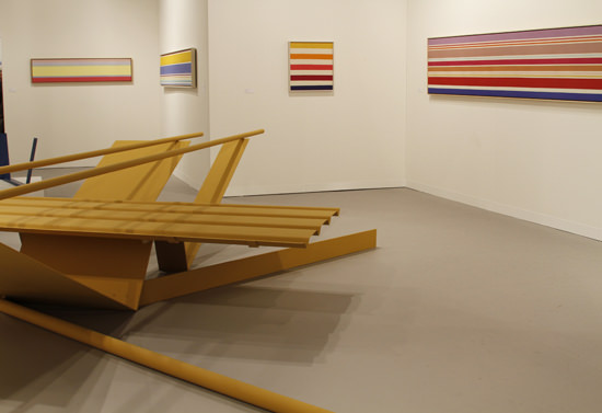 Installation photograph. An abstract sculpture made up of two long tubes balanced across V-shaped metal panels, all painted harvest yellow, is displayed in a gallery hung with horizontal, striped paintings.