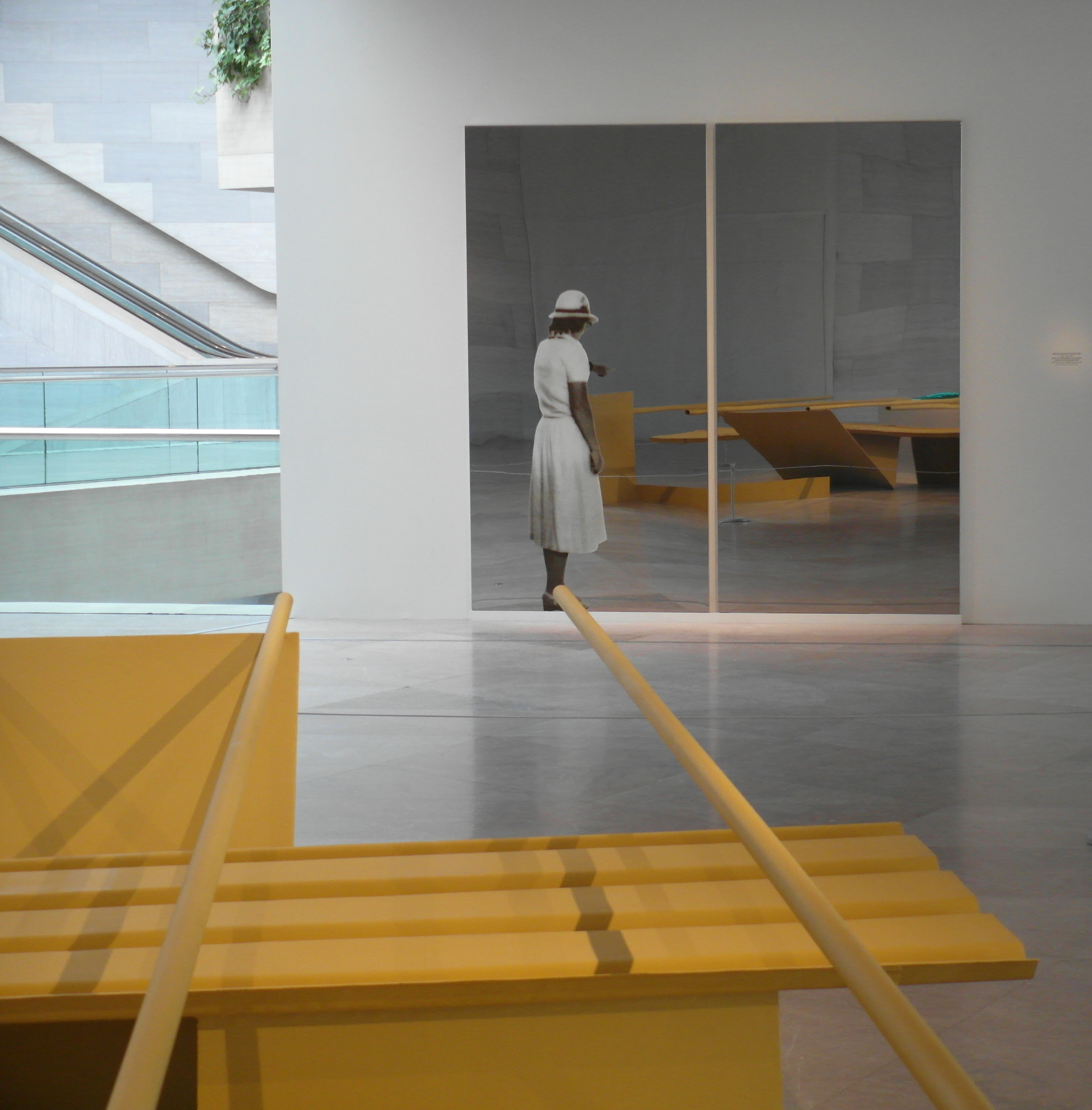 Installation photograph. The yellow tubes of an abstract sculpture angle towards a work of art made of two rectangular pieces of reflective metal hanging on the wall opposite us. A woman wearing a white dress and pointing is screen printed onto the left of two reflective panels. The abstract sculpture is also partially visible in the reflection.