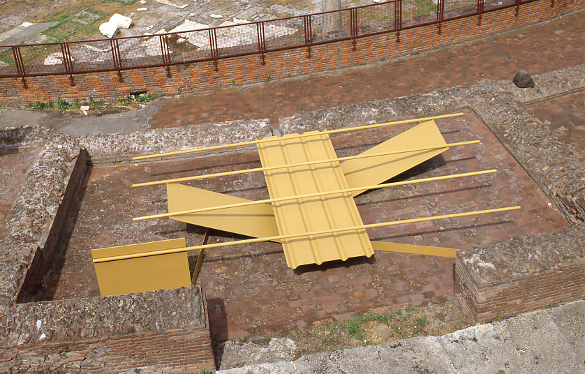 Sculpture. We look down onto a stone courtyard where four metal panels and four tubes, all muted yellow, are balanced to create this abstract sculpture. The four tubes are spaced along a wide strip, which in turn rests on V-shaped strips.