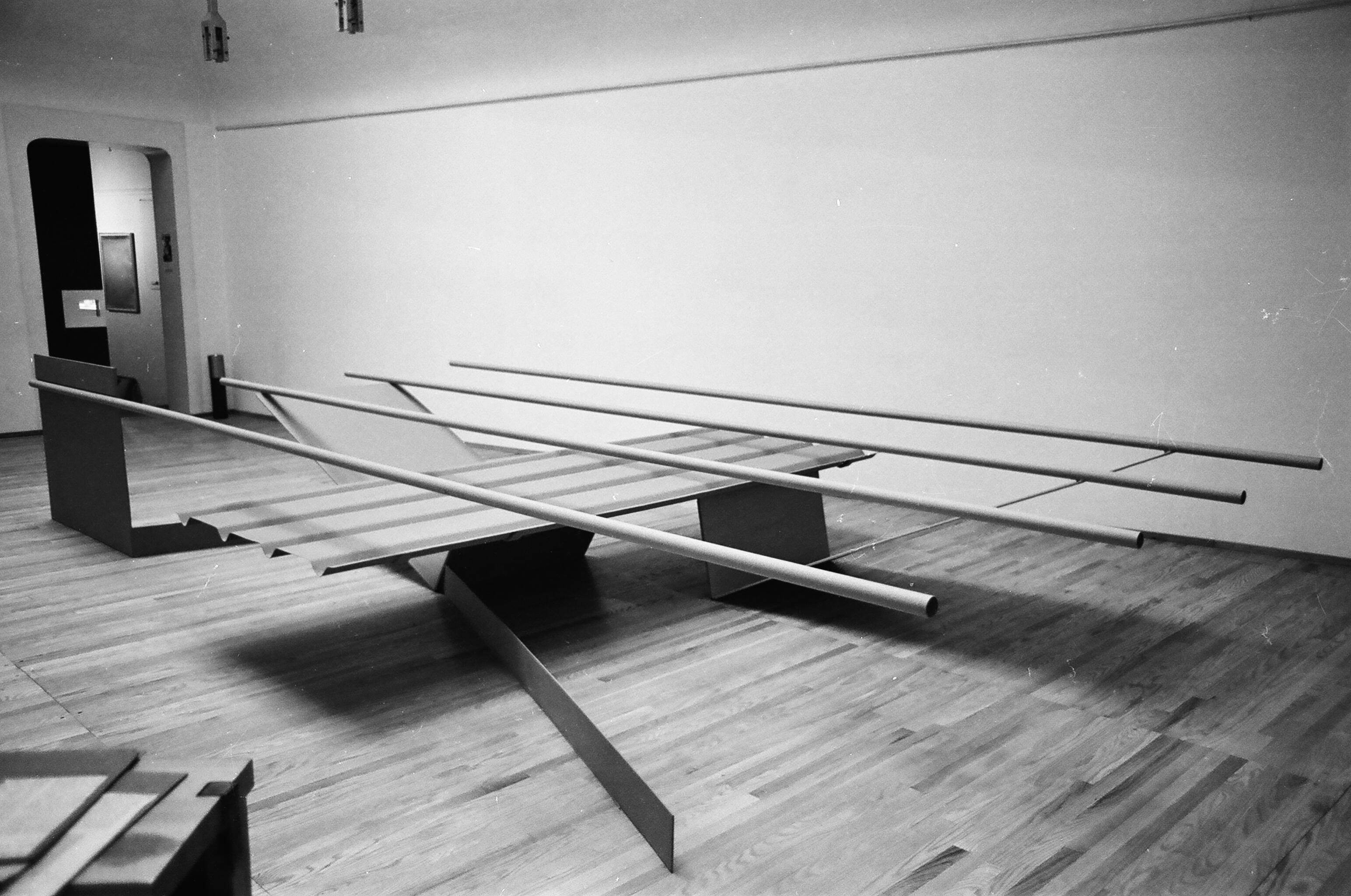 Installation photograph. Black and white photograph of an abstract sculpture made of four tubes balanced across four straight and V-shaped panels. The sculpture fills the part of the room we can see.