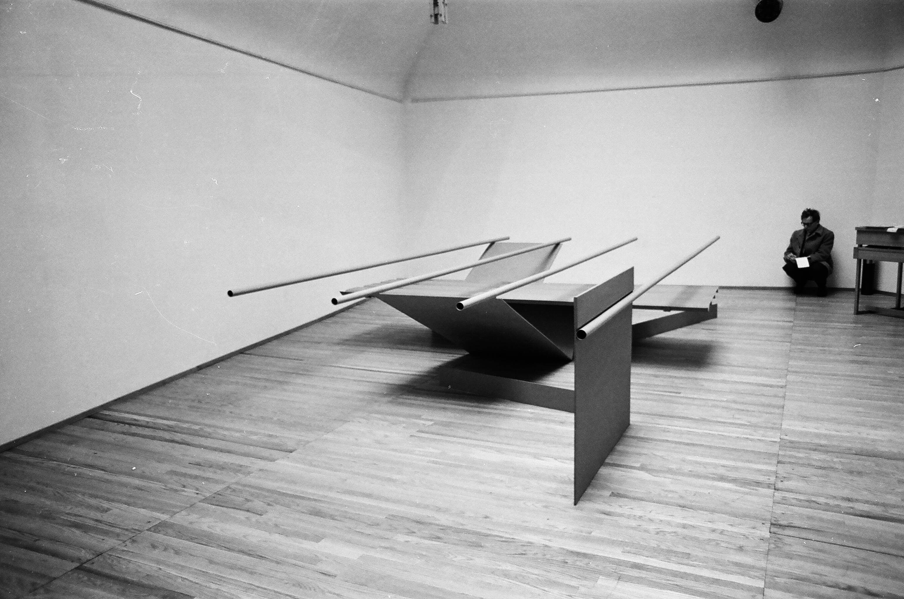 Installation photograph. Black and white photograph of an abstract sculpture. Four tubes attached to and balanced across vertical and V-shaped panels come towards us. A person crouches against the wall opposite us.