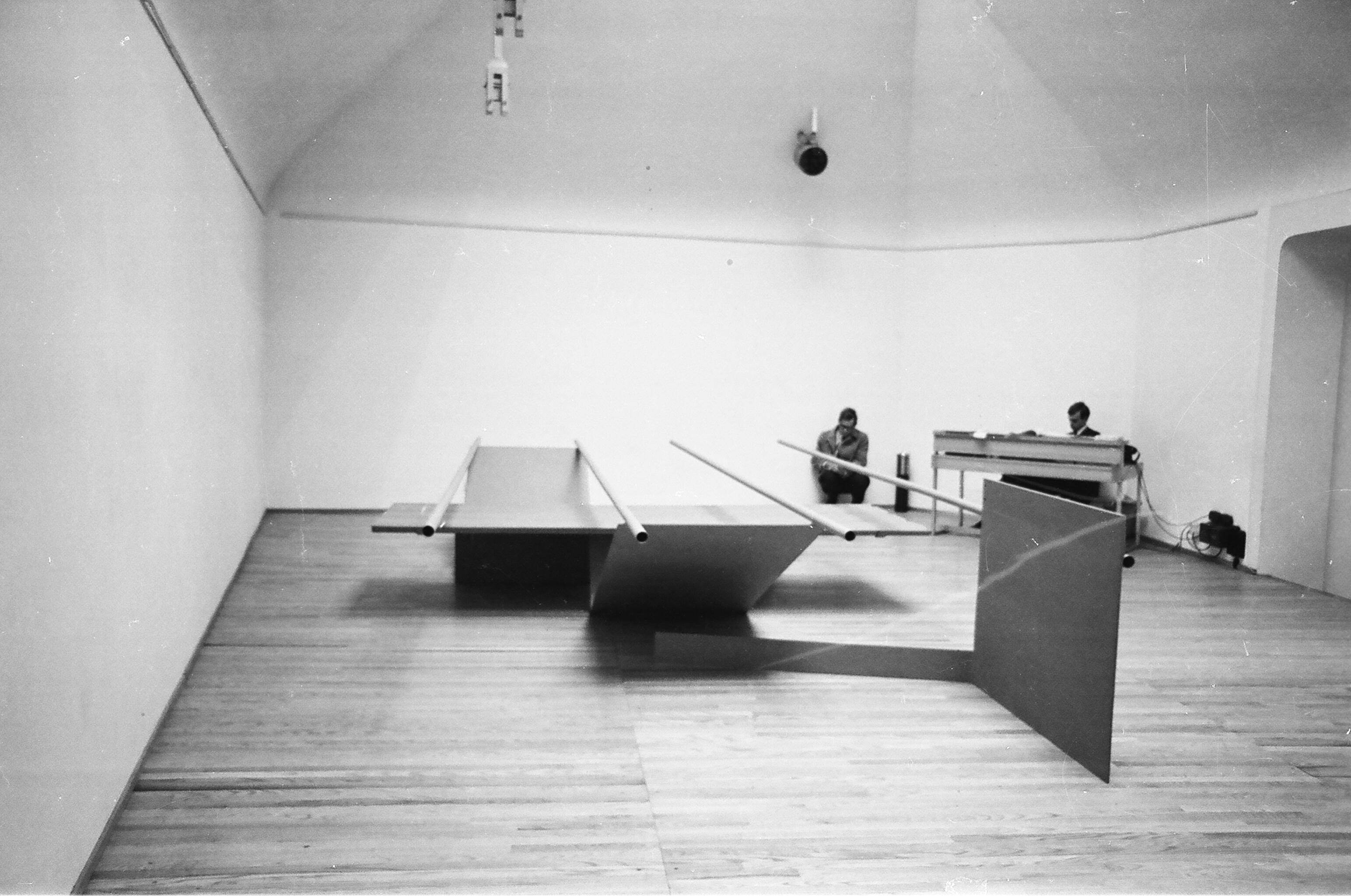 Installation photograph. Black and white photograph of an abstract sculpture. Four tubes balance across horizontal, vertical, and V-shaped metal panels. A person crouches against the wall opposite us, and another sits at a table in the corner.