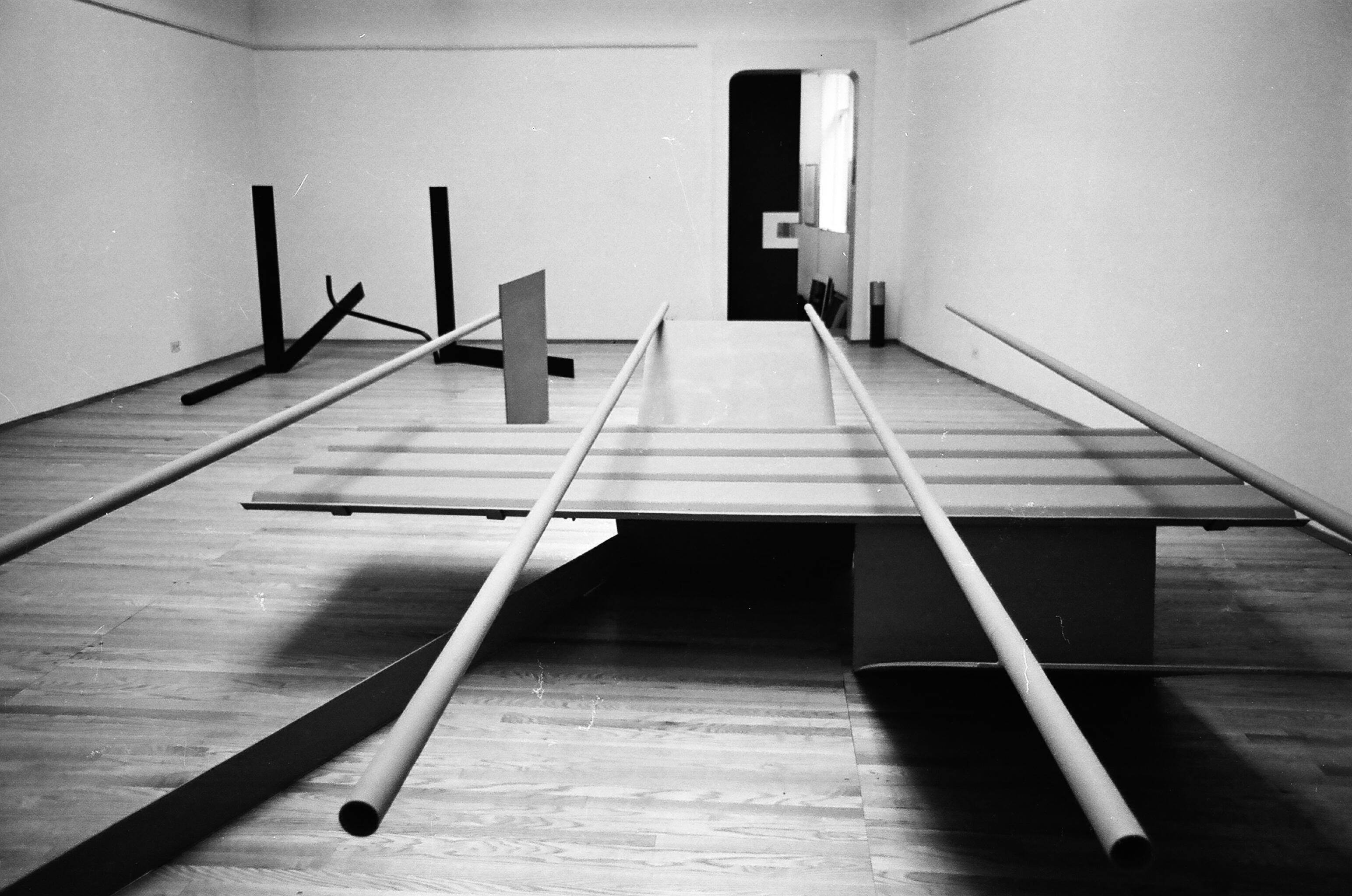 Installation photograph. Black and white photograph of an abstract sculpture. Four tubes balanced across flat and V-shaped metal panels come right at us like railroad tracks.