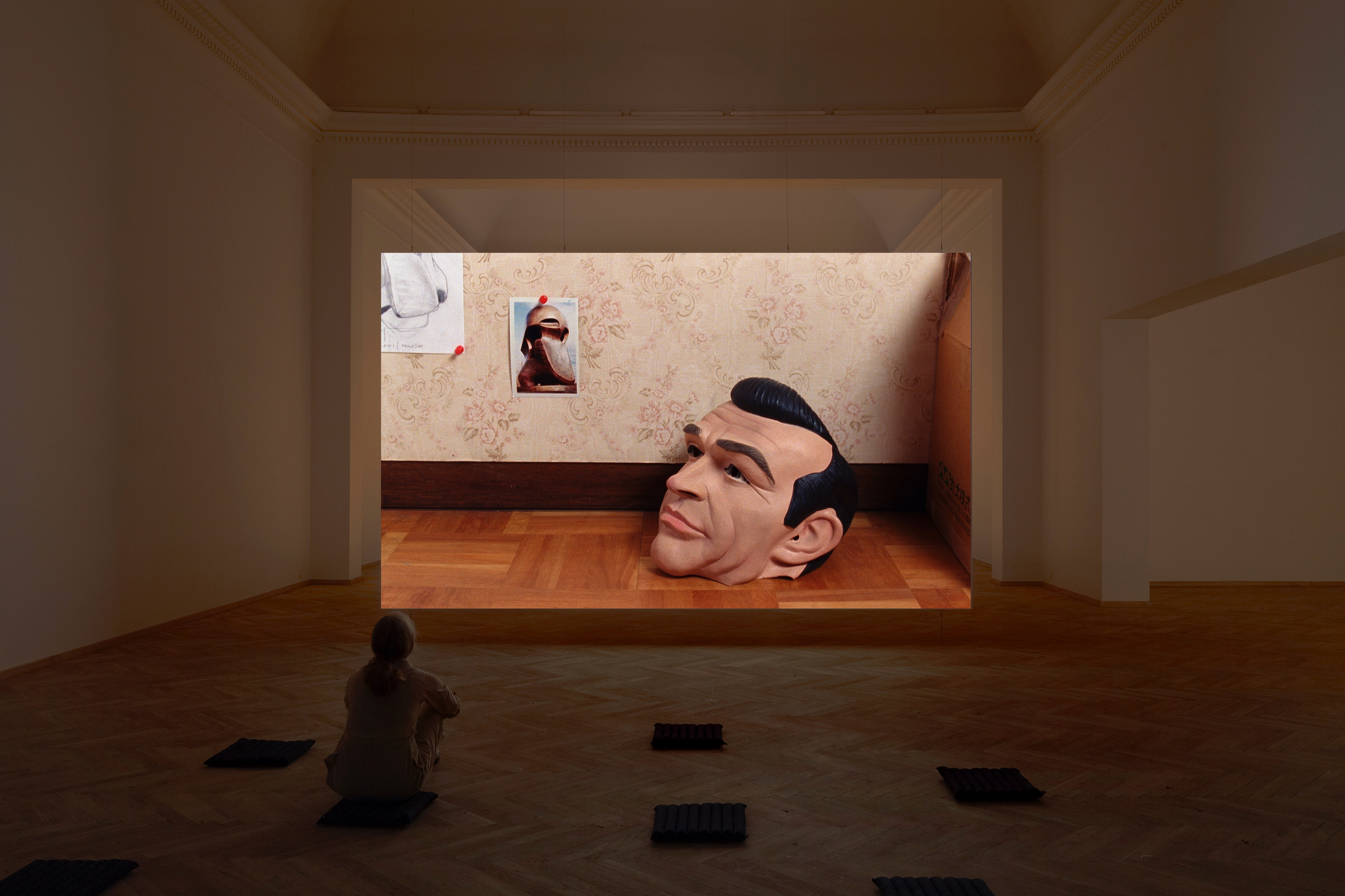 Installation photograph. A film is projected at the far end of a dim gallery. The still shows a full-head mask of Sean Connery with black hair. A person sits on a mat in the gallery, arms around bent knees.